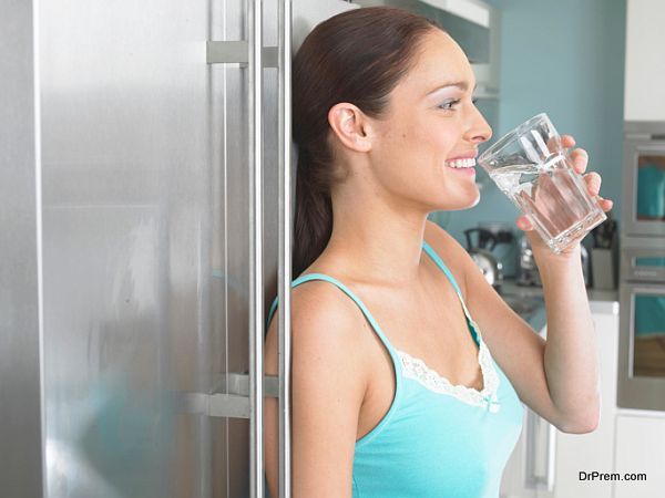 Young woman leaning against fridge, drinking glass of water, side view