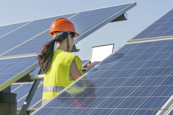 A woman in orange helmet is standing beside the solar energy panel and using the notebook